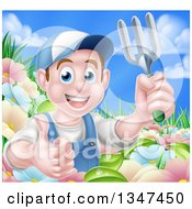 Happy Young Brunette White Male Gardener In Blue Holding Up A Garden Fork And Thumb In A Flower Garden