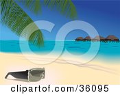 Pair Of Sunglasses Resting Under A Palm Tree On A Tropical Sandy Beach With Huts On The Water In The Background